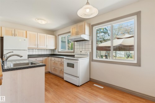 Kitchen featuring wood-finish flooring, light wood cabinetry, dark countertops, a white tile backsplash, and a double basin stainless steel sink - 10620 68 Avenue, Edmonton, AB - Indoor Photo Showing Kitchen