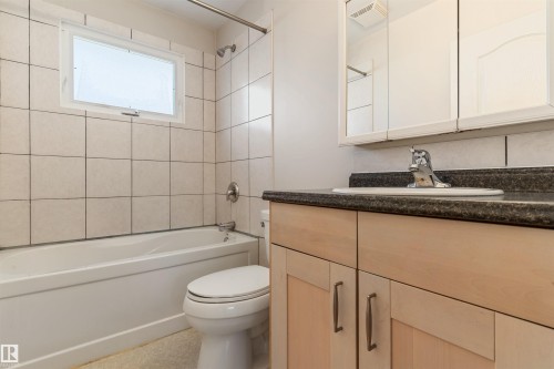 Full bathroom featuring a white bathtub with a shower fixture, light-toned wall tiling, a white toilet, and a vanity with a dark countertop and light wood-finish cabinetry - 10620 68 Avenue, Edmonton, AB - Indoor Photo Showing Bathroom