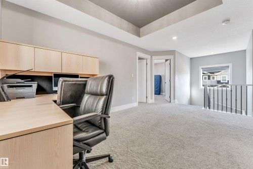 Loft area featuring a built-in wood-finish desk with upper cabinetry, tray ceiling detail, soft gray carpeting, and an open stairwell with black metal spindles - 1103 29 Street, Edmonton, AB - Indoor Photo Showing Office