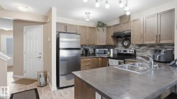 Modern kitchen featuring a dual basin sink with gooseneck faucet, light wood-finish cabinetry, stainless steel refrigerator, and dark countertop surfaces - 