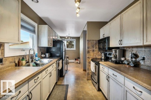 Galley kitchen featuring light-toned cabinetry, dark countertops, tiled backsplash, stainless steel sink, and tile flooring - 10616 136 Street, Edmonton, AB - Indoor Photo Showing Kitchen With Double Sink