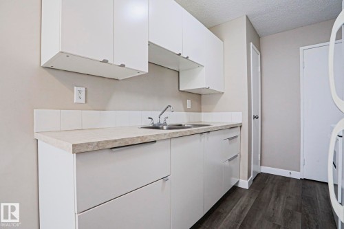 Kitchenette featuring white cabinetry, light-toned countertops, a stainless steel double sink, a white tile backsplash, and wood-finish flooring - 118 7220 144 Avenue, Edmonton, AB - Indoor