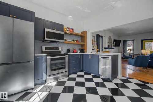 Modern kitchen featuring checkerboard flooring, navy cabinetry with gold-tone hardware, stainless steel appliances, open shelving, and a tile backsplash - 11512 122 St, Edmonton, AB - Indoor Photo Showing Kitchen