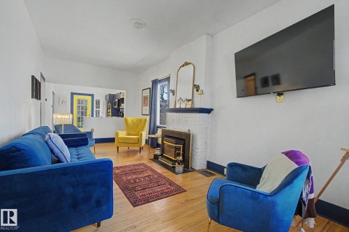 Living area featuring wood-finish flooring, a white brick fireplace with a dark mantel, a large wall-mounted display, and a half-wall partition - 11512 122 St, Edmonton, AB - Indoor Photo Showing Living Room