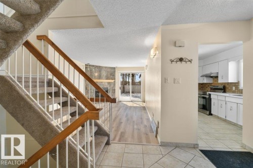 The property features a staircase with carpeting and wooden handrails, a living area with hard surface flooring and a stone-clad fireplace, and a kitchen with white cabinetry and a tiled backsplash - 17313 60 Avenue, Edmonton, AB - Indoor Photo Showing Other Room