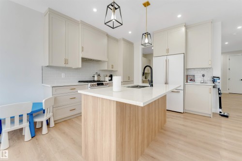 Spacious kitchen featuring an island with wood-finish paneling and a light-toned countertop, white shaker-style cabinetry with gold-tone hardware, a subway tile backsplash, and light wood-finish flooring - 7007 169 Avenue Nw, Edmonton, AB - Indoor Photo Showing Kitchen With Double Sink With Upgraded Kitchen