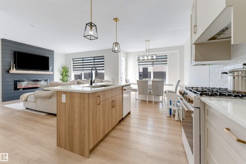 Open-concept living space featuring wood-finish flooring, a kitchen island with integrated sink, stainless steel range, and a living area with a dark paneled accent wall and linear electric fireplace - 7007 169 Avenue Nw, Edmonton, AB - Indoor Photo Showing Kitchen With Upgraded Kitchen