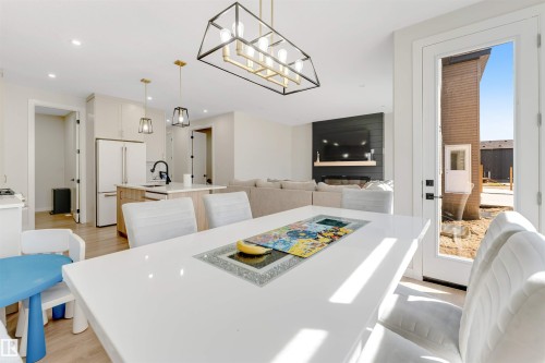 Open-concept living space with wood-finish flooring, a contemporary kitchen featuring light-toned cabinetry and an island, a dining area with a high-gloss table and upholstered chairs, and a living room with a shiplap accent wall and fireplace - 7007 169 Avenue Nw, Edmonton, AB - Indoor Photo Showing Dining Room