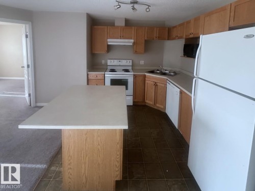 Kitchen featuring a central island with an extended countertop, wood-finish cabinetry, a white electric range, a double basin stainless steel sink, and white appliances - 6120 7331 South Terwillegar Drive Nw, Edmonton, AB - Indoor Photo Showing Kitchen With Double Sink