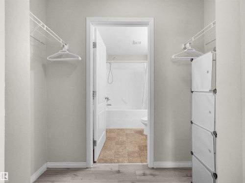 Walk-through closet featuring wire shelving and wood-finish flooring, opening to a bathroom with a white tub-shower combination and tiled flooring - 7123 7327 South Terwillegar Drive, Edmonton, AB - Indoor With Storage
