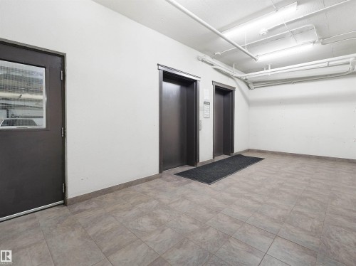 Tile flooring throughout the common area, featuring two elevators with brushed metal doors and a fire-rated door with a wired glass window - 7123 7327 South Terwillegar Drive, Edmonton, AB - Indoor Photo Showing Other Room