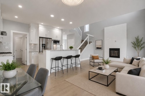 Open-concept living space featuring a gourmet kitchen, integrated breakfast bar, tall fireplace, light wood-finish flooring, and a modern staircase with glass railing. Virtually Staged. - 635 42 Street, Edmonton, AB - Indoor
