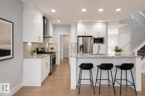 Modern kitchen featuring white cabinetry, stainless steel appliances, light wood-finish flooring, a stone-finish countertop island with seating, and recessed lighting. Virtually Staged. - 635 42 Street, Edmonton, AB - Indoor Photo Showing Kitchen With Upgraded Kitchen