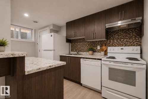 Contemporary kitchen featuring dark wood-finish cabinetry, light-toned countertops, a mosaic tile backsplash, stainless steel sink, and white appliances. Virtually Staged. - 635 42 Street, Edmonton, AB - Indoor Photo Showing Kitchen