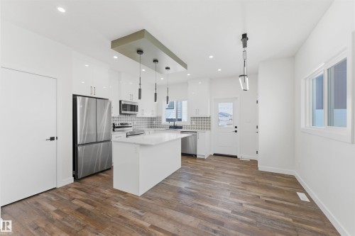 Bright kitchen featuring an island with a light-toned countertop, stainless steel appliances, a patterned tile backsplash, and sleek white cabinetry - 12447 47 Street, Edmonton, AB - Indoor Photo Showing Kitchen With Stainless Steel Kitchen With Upgraded Kitchen