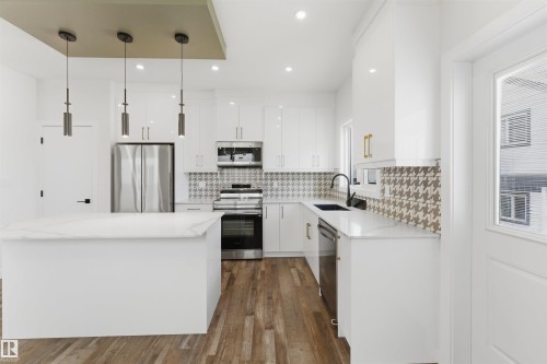 Modern kitchen featuring white cabinetry, a large center island with a light-toned countertop, stainless steel appliances, wood-finish flooring, and a patterned backsplash - 12447 47 Street, Edmonton, AB - Indoor Photo Showing Kitchen With Stainless Steel Kitchen With Upgraded Kitchen