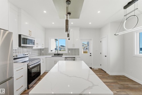 Bright kitchen featuring a large central island with a stone-finish countertop, white cabinetry with gold-tone hardware, stainless steel appliances, and wood-finish flooring - 12447 47 Street, Edmonton, AB - Indoor Photo Showing Kitchen With Stainless Steel Kitchen With Upgraded Kitchen