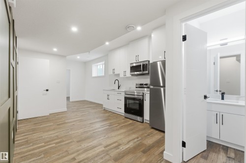 Open-concept living space featuring wood-finish flooring, recessed lighting, and a kitchen with stainless steel appliances, white cabinetry, and matte black fixtures - 12447 47 Street, Edmonton, AB - Indoor Photo Showing Kitchen With Stainless Steel Kitchen