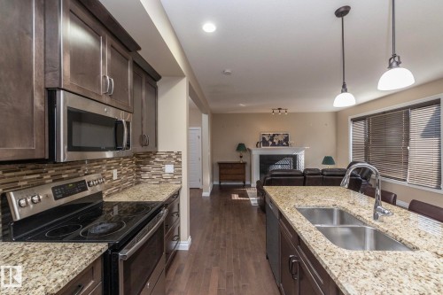 Open-concept kitchen featuring granite countertops, dark wood cabinetry, stainless steel appliances, a mosaic tile backsplash, and wood-finish flooring - 151 Caledonia Drive, Leduc, AB - Indoor Photo Showing Kitchen With Double Sink With Upgraded Kitchen