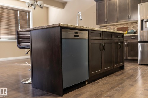 Kitchen island featuring a dual-basin sink, granite countertop, integrated dishwasher, and dark wood cabinetry - 151 Caledonia Drive, Leduc, AB - Indoor Photo Showing Kitchen