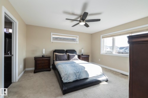 Carpeted room with a ceiling fan, two windows with blinds, and a doorway revealing a partial bathroom vanity - 151 Caledonia Drive, Leduc, AB - Indoor Photo Showing Bedroom