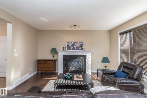 Living area featuring a fireplace with a black surround and white mantel, track lighting, wood-finish flooring, and a large window with blinds - 151 Caledonia Drive, Leduc, AB - Indoor Photo Showing Living Room With Fireplace