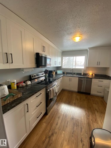 The kitchen features white cabinetry with dark hardware, dark countertops, stainless steel appliances, and wood-style flooring - 5030 40 Street, Cold Lake, AB - Indoor Photo Showing Kitchen