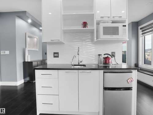 Compact kitchen island featuring white cabinetry with brushed nickel pulls, a dark countertop, an integrated sink with a gooseneck faucet, a built-in microwave, and a compact stainless steel refrigerator - 913 Wood Place, Edmonton, AB - Indoor Photo Showing Kitchen With Upgraded Kitchen