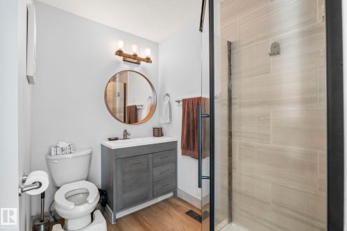 Bathroom featuring a glass-enclosed shower with tiled walls, a vanity with a white countertop and gray cabinetry, wood-finish flooring, a round mirror, and a three-light fixture - 23133 Twp Rd 502, Rural Leduc County, AB - Indoor Photo Showing Bathroom