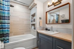 Bathroom featuring a white tub with light-toned tiled surround, built-in shower niches, a vanity with a light wood-finish countertop, and a vessel sink with a chrome faucet - 