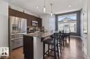 Open-concept kitchen and dining area featuring dark wood-finish flooring, stainless steel appliances, a breakfast bar with stone-finish countertop, dark cabinetry, and an expansive window arrangement - 17540 58 Street Nw, Edmonton, AB 