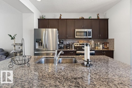 Kitchen featuring a granite-finish island with an undermount double basin sink, stainless steel appliances, dark wood-finish cabinetry, and a subway tile backsplash - 17540 58 Street Nw, Edmonton, AB 