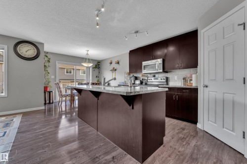 Open-concept kitchen featuring an expansive center island with a light-colored countertop, dark wood-finish cabinetry, stainless steel appliances, and wood-finish flooring - 5520 5 Avenue, Edmonton, AB - Indoor Photo Showing Kitchen With Upgraded Kitchen