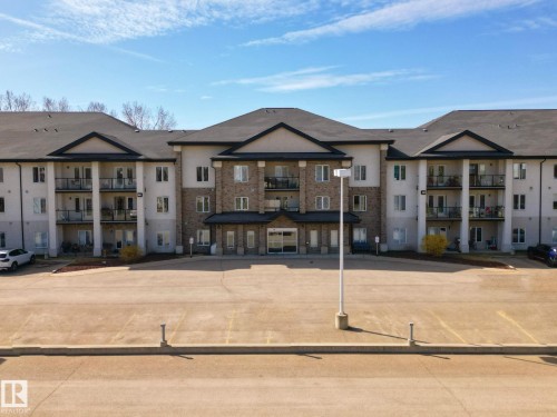 Multi-story building featuring a brick and stucco facade, individual balconies with glass railings, a central covered entrance, and a paved parking lot - 220 80A Kelloway Crescent, Red Deer, AB - Outdoor With Balcony With Facade