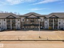 Multi-story building featuring a brick and stucco facade, individual balconies with glass railings, a central covered entrance, and a paved parking lot - 220 80A Kelloway Crescent, Red Deer, AB  - Outdoor With Balcony With Facade 