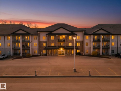 Multi-story residential building featuring a stucco and stone facade, multiple private balconies, a covered main entrance, and an expansive paved parking area - 220 80A Kelloway Crescent, Red Deer, AB - Outdoor With Balcony