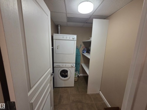 Dedicated laundry area featuring a stacked washer and dryer unit, tile flooring, built-in shelving, and neutral wall tones - 109 160 Magrath Rd, Edmonton, AB - Indoor Photo Showing Laundry Room