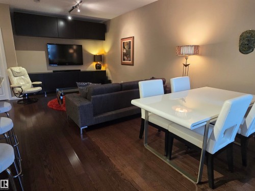 Open-concept living and dining area featuring dark wood-finish flooring, a contemporary white dining table, built-in media cabinetry, and track lighting - 109 160 Magrath Rd, Edmonton, AB - Indoor