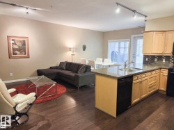 Open-concept living area with wood-finish flooring, a kitchen peninsula featuring a granite-style countertop, light wood cabinetry, and track lighting - 
