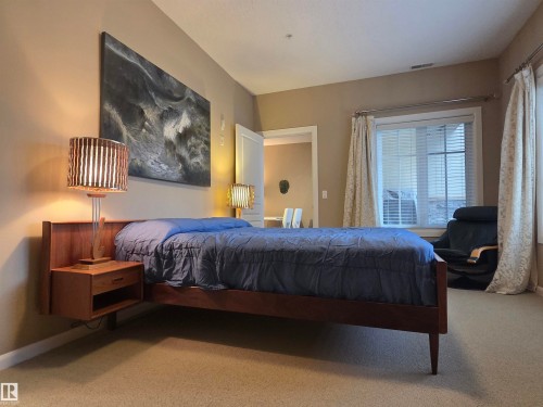 Carpeted bedroom featuring a window with blinds and drapes, a wood-finish bed frame, and a built-in floating nightstand - 109 160 Magrath Rd, Edmonton, AB - Indoor Photo Showing Bedroom