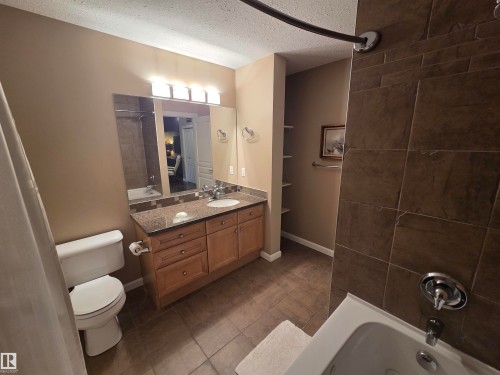 Bathroom featuring a light wood-finish vanity with a speckled countertop and integrated sink, a rectangular mirror, and an overhead vanity light fixture - 109 160 Magrath Rd, Edmonton, AB - Indoor Photo Showing Bathroom