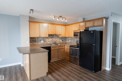 Kitchen featuring light wood cabinetry, a mosaic tile backsplash, dark countertops, and wood-finish flooring - 4407 23 Street, Edmonton, AB - Indoor Photo Showing Kitchen With Double Sink
