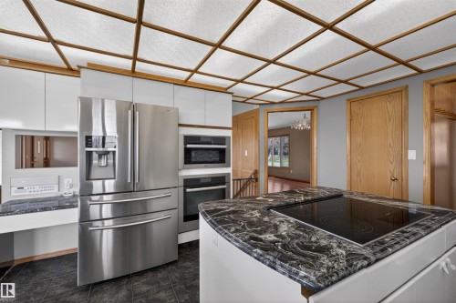 Kitchen featuring an island with a built-in cooktop and dark patterned countertop, stainless steel appliances, white cabinetry, and a grid-patterned ceiling with translucent panels - 19403 Fort Road, Edmonton, AB - Indoor Photo Showing Kitchen