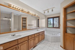Bathroom featuring a double vanity with wood cabinetry and white countertops, a large framed mirror with overhead lighting, a built-in corner bathtub with white tile surround, and a separate glass-enclosed shower - 