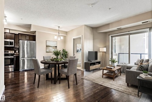 Open-concept living space featuring wood-finish flooring, a kitchen with dark cabinetry and stainless steel appliances, a dining area with a chandelier, and a living area with a sliding glass door to a balcony - 316 10520 56 Avenue Nw, Edmonton, AB - Indoor