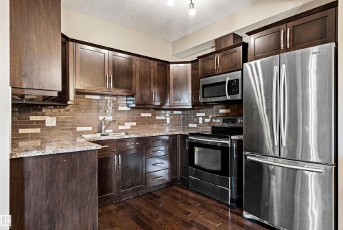 Kitchen featuring dark wood cabinetry, stainless steel appliances, a mosaic tile backsplash, wood-finish flooring, and light-toned countertops - 316 10520 56 Avenue Nw, Edmonton, AB - Indoor Photo Showing Kitchen With Stainless Steel Kitchen With Double Sink