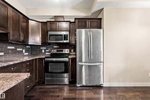 Kitchen featuring dark wood cabinetry, stainless steel appliances, a mosaic tile backsplash, and wood-finish flooring - 316 10520 56 Avenue Nw, Edmonton, AB - Indoor Photo Showing Kitchen With Stainless Steel Kitchen