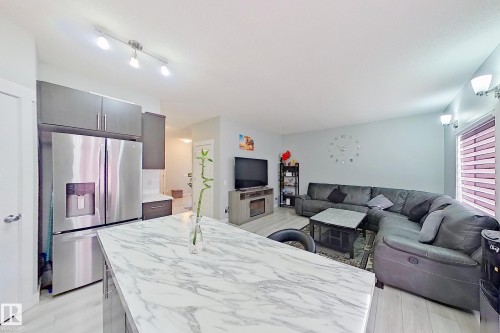 Open-concept living space featuring a kitchen island with a marble-look countertop, stainless steel refrigerator, dark wood-finish cabinetry, light wood-finish flooring, and track lighting - 1221 27 Street, Edmonton, AB - Indoor