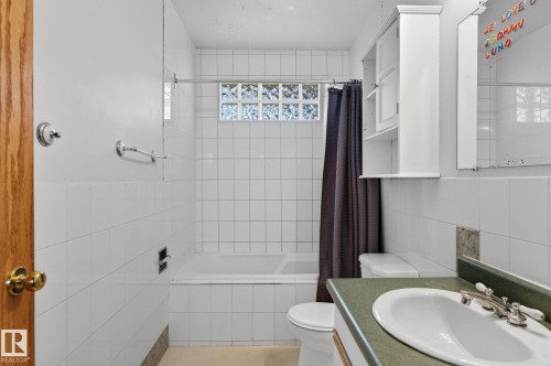 Bathroom featuring a white tile surround bathtub with glass block window, a white toilet, and a vanity with an integrated sink and green countertop - 13412 108 Street, Edmonton, AB - Indoor Photo Showing Bathroom