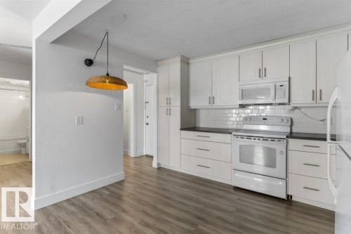 Kitchen featuring light wood-finish flooring, white cabinetry, dark countertops, a white subway tile backsplash, and white appliances - 101 8604 Gateway Boulevard, Edmonton, AB - Indoor Photo Showing Kitchen